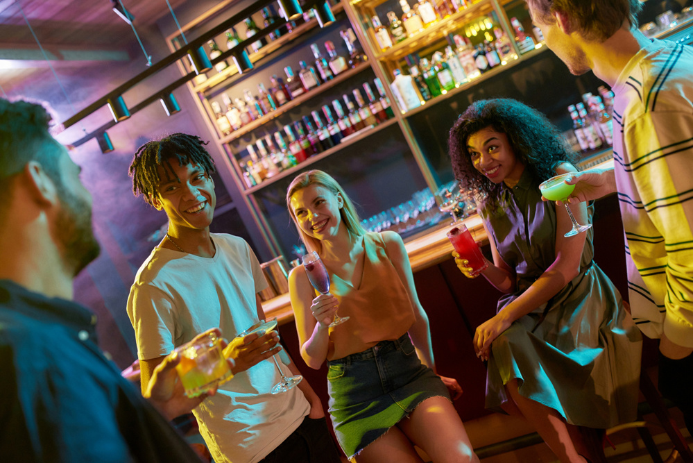 Nightlife. Cheerful young men drinking cocktail at the bar counter and flirting with women they just met. Young adults having drinks while chatting at the night club