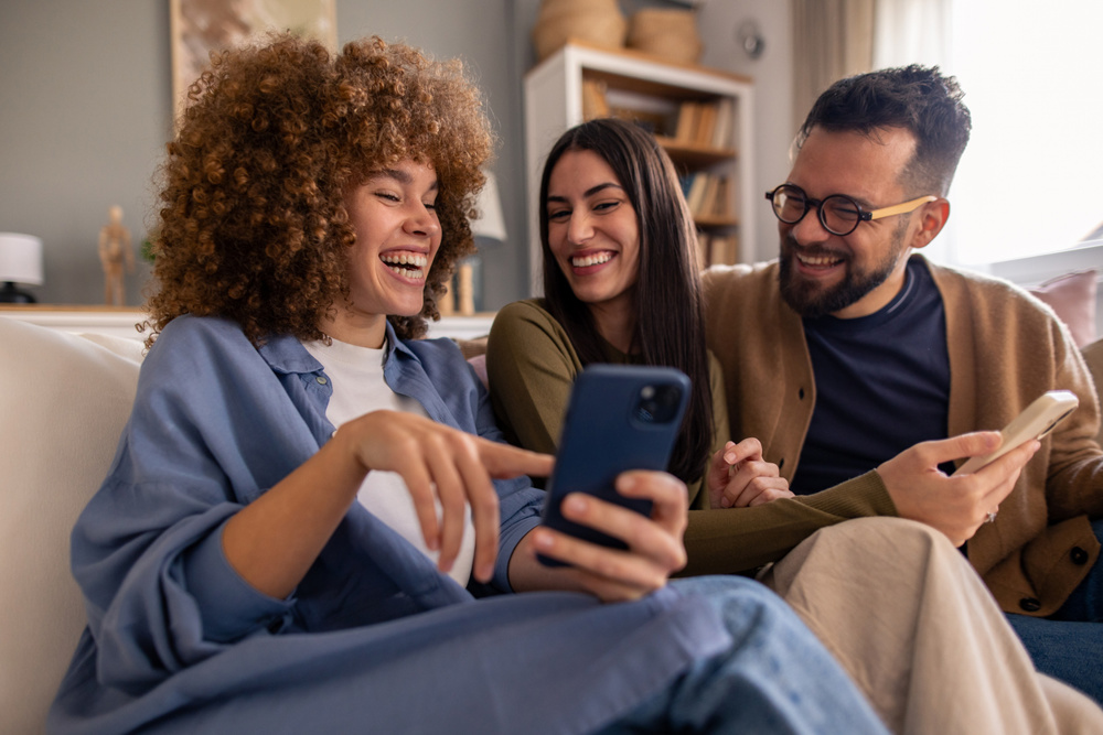 Diverse Friends Laughing Together While Sharing Social Media Content On Cozy Living Room Sofa