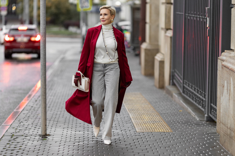 Stylish Woman in Burgundy Coat Walks City Street. Wearing Fashionable clothing White Turtleneck Sweater, Pearl Necklace and Grey Jeans, Carrying White Bag