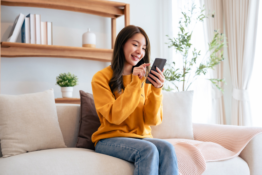 Happy young asian woman relax on comfortable couch at home texting messaging on smartphone, smiling girl use cellphone chatting, browse wireless internet on gadget