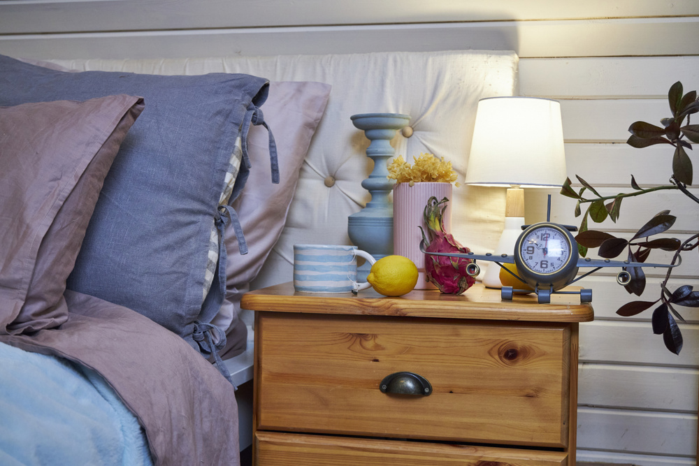 A fragment of a modern bedroom interior in a wooden house, featuring a wooden nightstand with bright decor next to a bed with blue and pink bedding