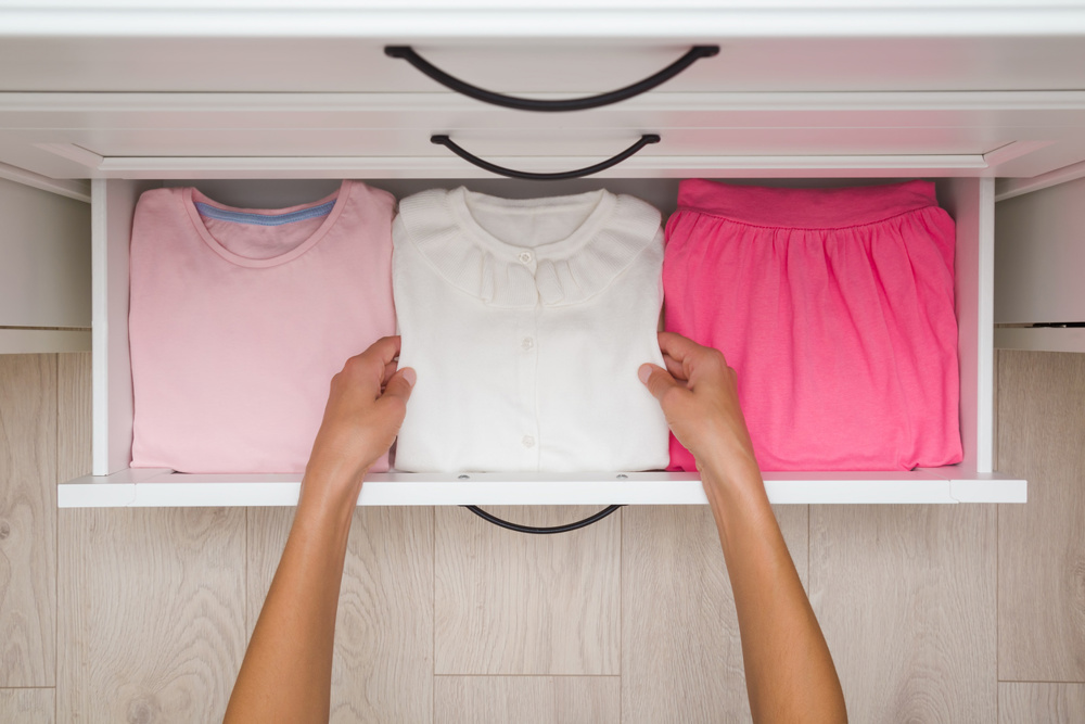 Young mother hands sorting pink white folded knitted sweater, t shirt and skirt in opened drawer box at home room. Making neatness. Child girl clothes. Closeup. Point of view shot. Top down view
