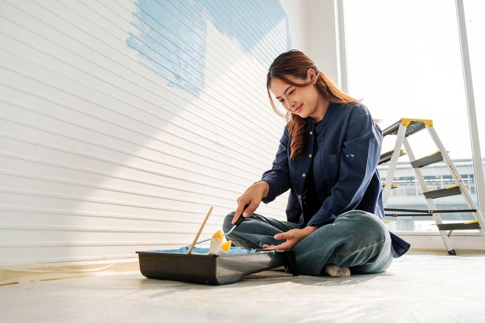 Smiling woman prepares blue paint during a DIY home renovation project. Sitting on the floor with tools and a step ladder, she embraces creativity and improvement in a naturally lit modern space