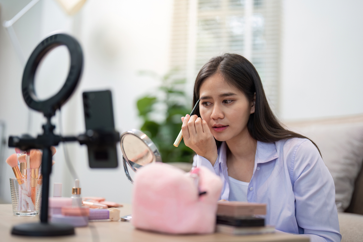 Asian woman concentrating while refining her eyebrow makeup, utilizing a mirror amid her home beauty routine setup