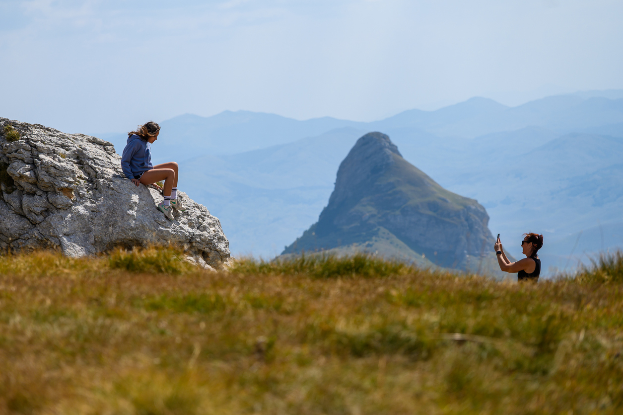 A woman takes a smartphone photo of a girl sitting on a rock at Sedlo Pass in Durmitor National Park, Montenegro, with the dramatic Saddle mountain peak in the hazy blue background