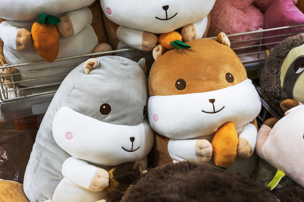Close-up of soft grey and brown hamster stuffed animals holding small plush carrots and acorns. The toys feature sweet smiling faces and are neatly arranged on a retail shelf, perfect for childhood and gift-themed content