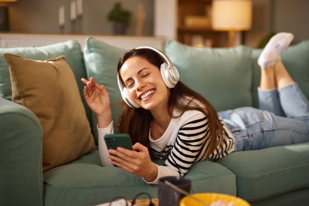 Young woman enjoying music through headphones, lying on the sofa and holding a smartphone