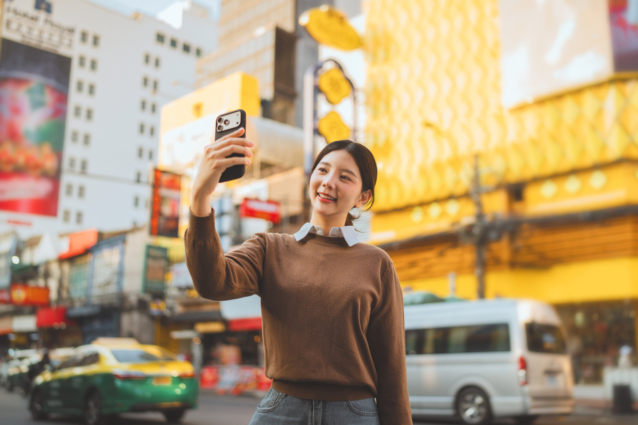 Happy Asian woman traveler taking selfie with smartphone at Yaowarat Road, young lady enjoying street food and neon lights at Bangkok Chinatown night market, concept of Thailand travel, urban lifestyle, and solo journey in famous landmark