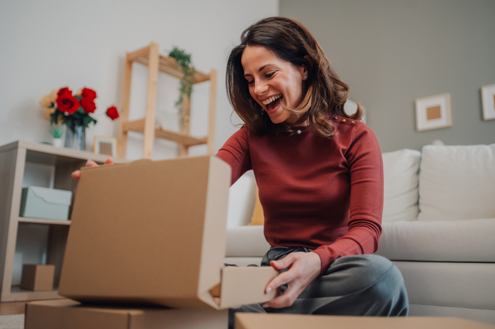 Excited woman opening a cardboard box in her living room, discovering a pleasant surprise