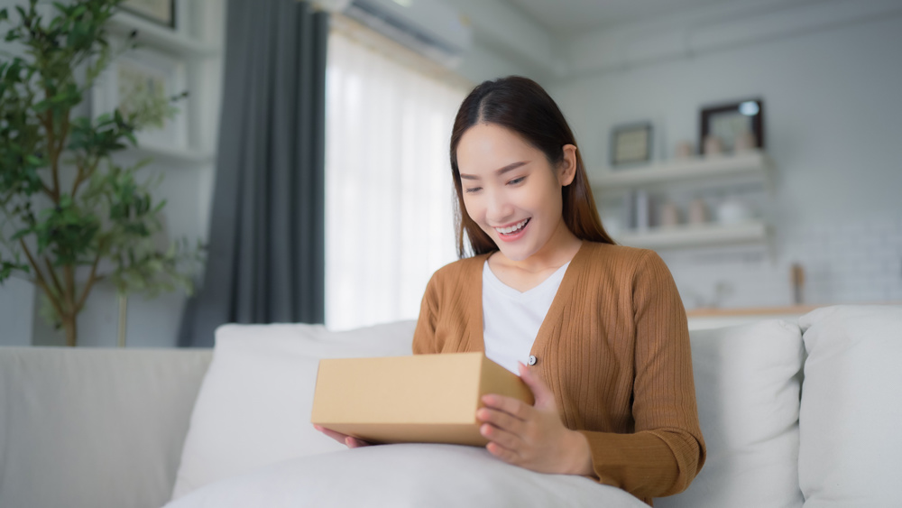 Happy young Asian woman opening delivery package at home. Beautiful female customer open box and sitting on couch in living room