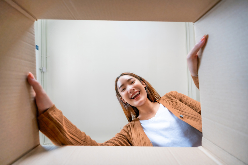 Low angle view of surprised young Asian woman unpacking. Opening carton box and looking inside. Packaging box, delivery service. Human emotions and facial expression